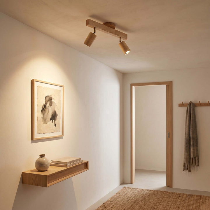 Two-head Merlo natural wood track lighting directing warm light toward framed art in a minimalist hallway