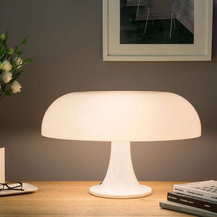 Glowing white Nesso Table Lamp on a wood desk with books and flowers