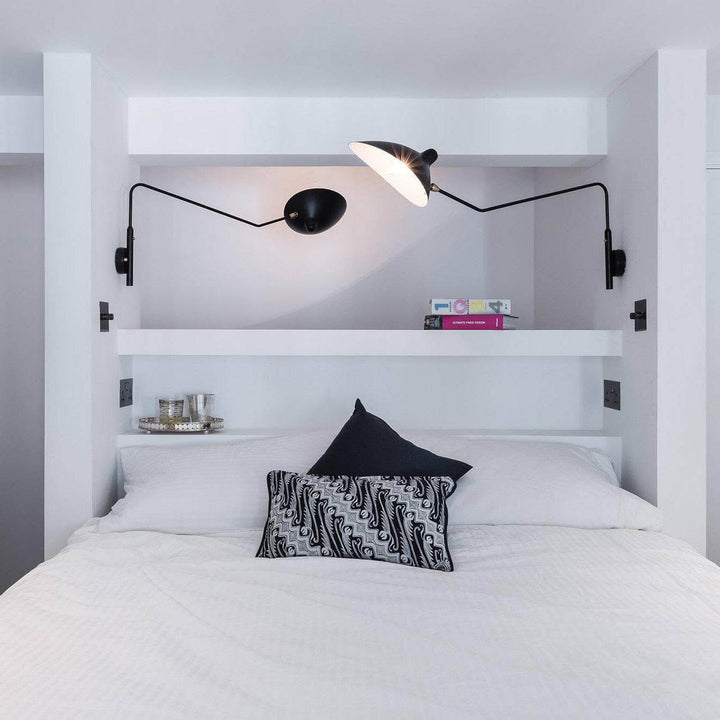 Two separate black swing arm wall sconces crossing above a white bedroom bed
