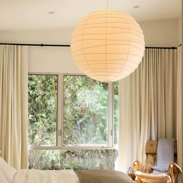 Large paper lantern pendant light over a bedroom with cream curtains and a window view.