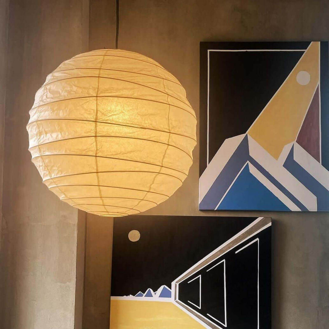 Round paper lantern pendant light glowing beside abstract wall art on a concrete wall.
