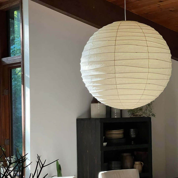 Round paper lantern pendant light near a tall window and black open shelving in a wood-ceiling room.