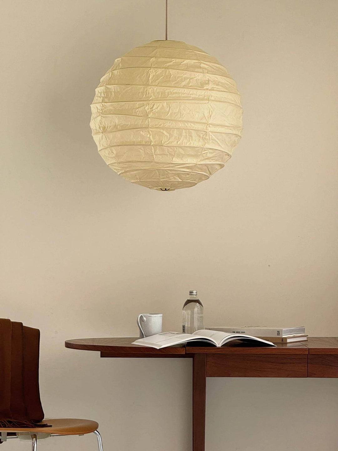 Paper lantern pendant light above a wood desk with books, cup, and plain wall in a quiet room.