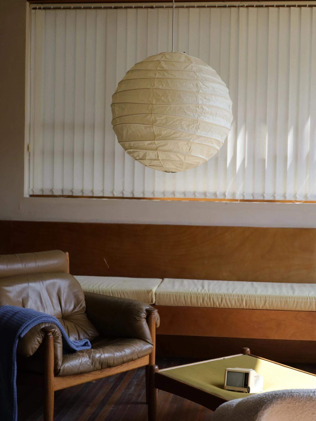 Round paper lantern pendant light in a lounge with vertical blinds, leather chair, and table.