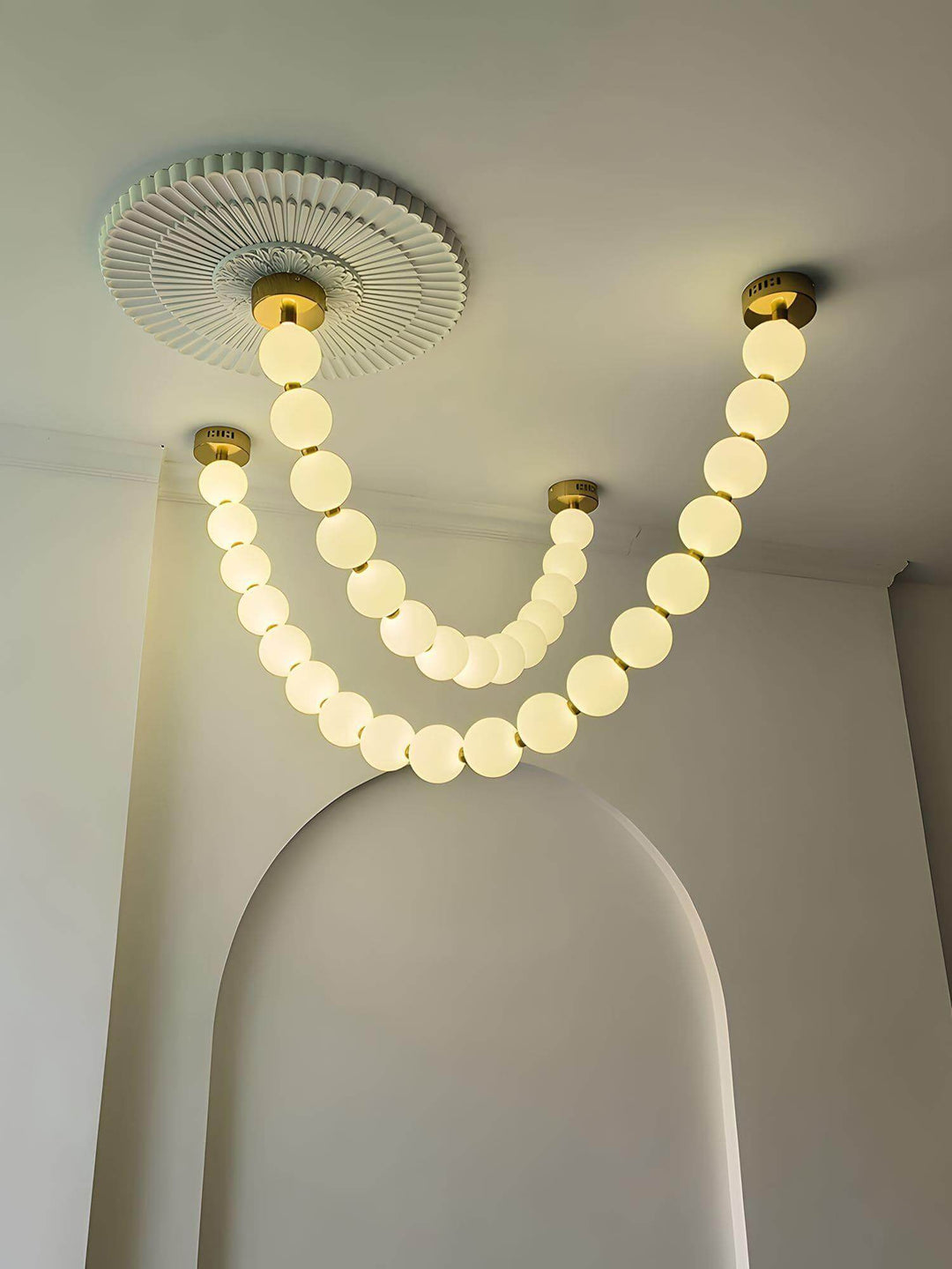 Warm glowing pearl necklace chandelier under a ceiling medallion near an arched wall.