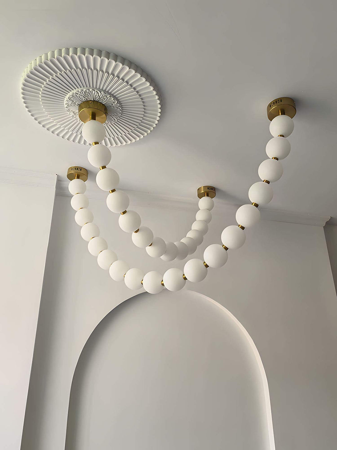 Unlit pearl necklace chandelier with white globes and brass accents near an arched wall.