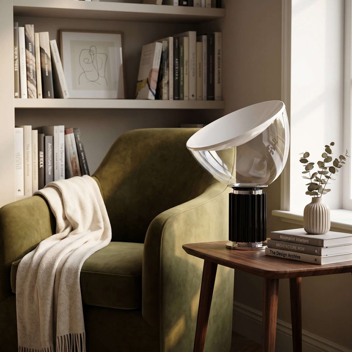 Black Taccia table lamp on a wood side table beside a green chair and bookshelf in daylight