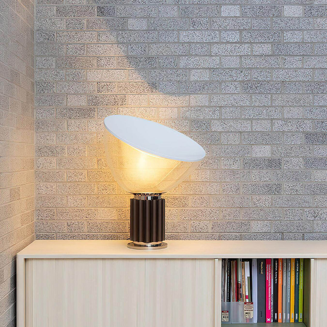 Glowing coffee-base Taccia table lamp on a white cabinet against a textured gray brick wall
