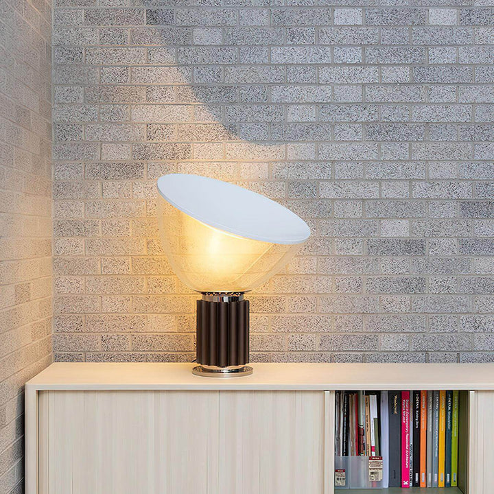 Glowing coffee-base Taccia table lamp on a white cabinet against a textured gray brick wall