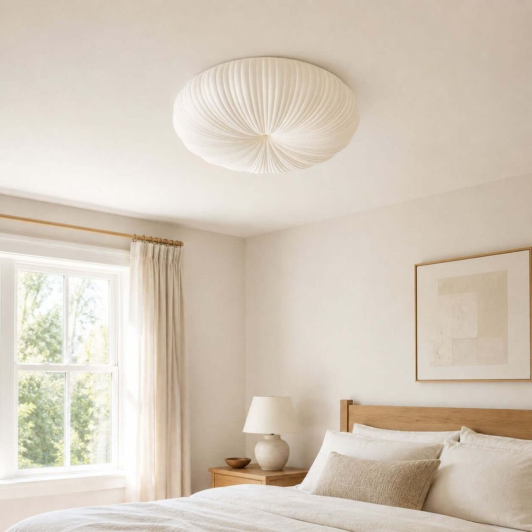 White pleated ceiling light shown close above a bed near a window.