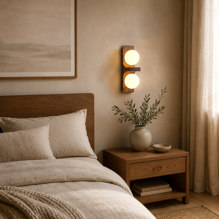 Double wall sconce glowing beside a bed and wooden nightstand in a warm bedroom.