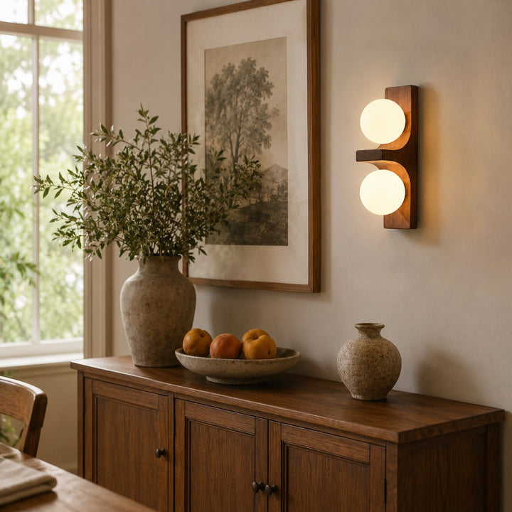 Wood double wall sconce glowing above a cabinet with framed art and decor.