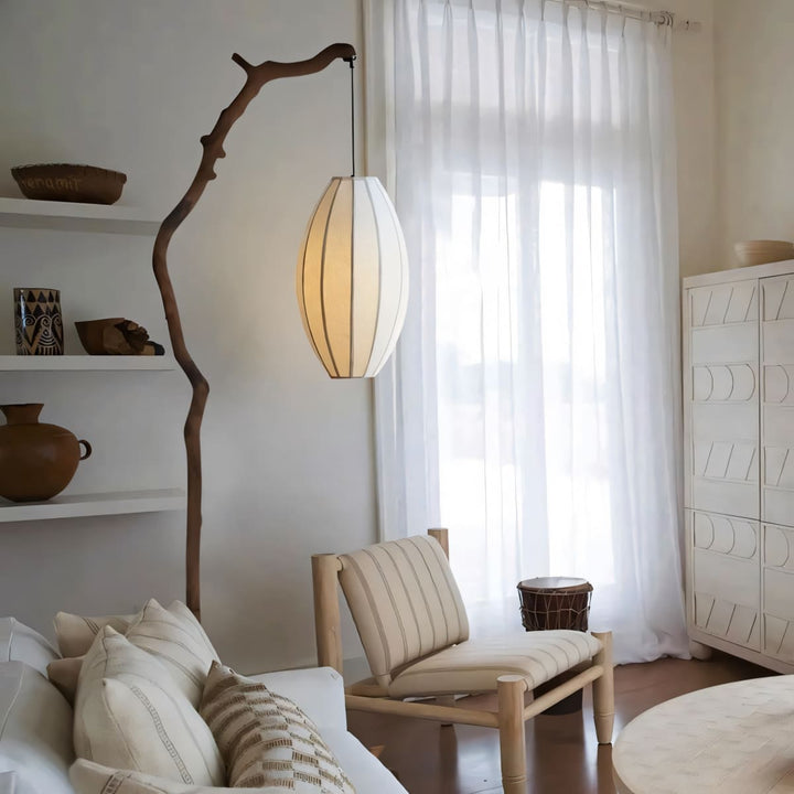 Tree branch floor lamp with fabric shade beside a lounge chair near sheer curtains.
