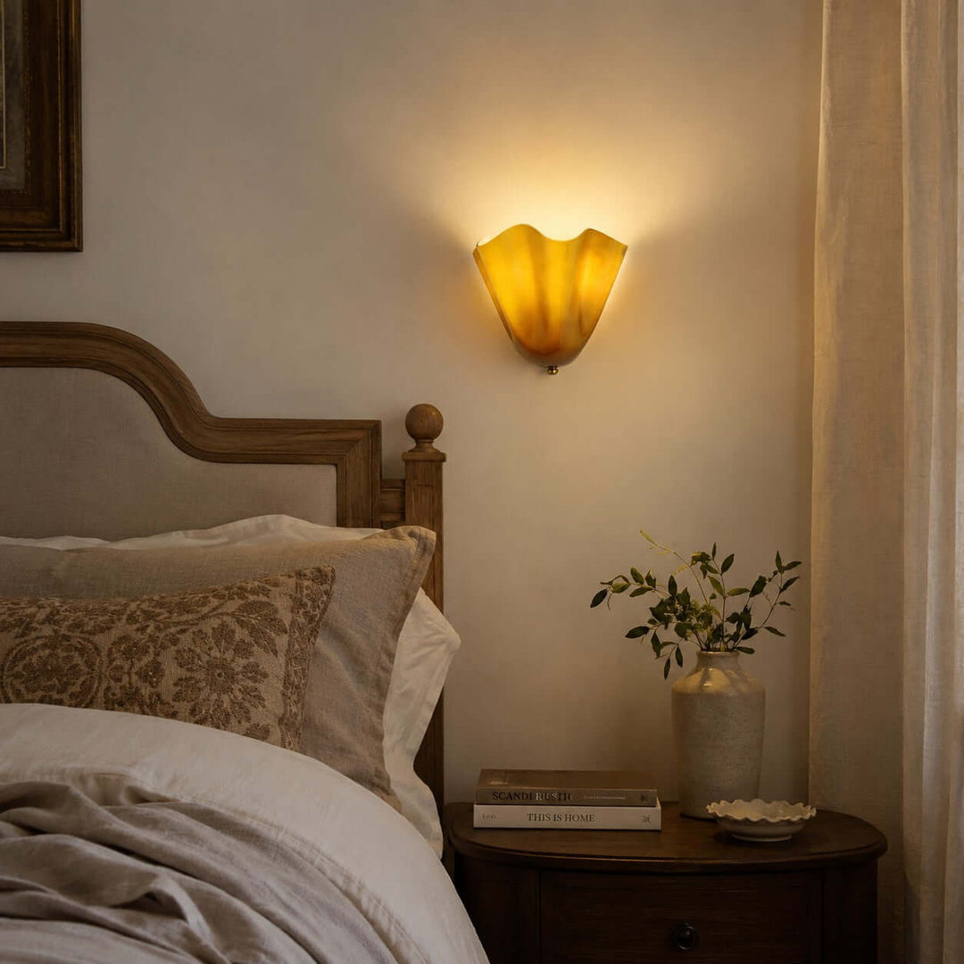 Amber glass wall sconce glowing beside a bed in a warm bedroom scene.