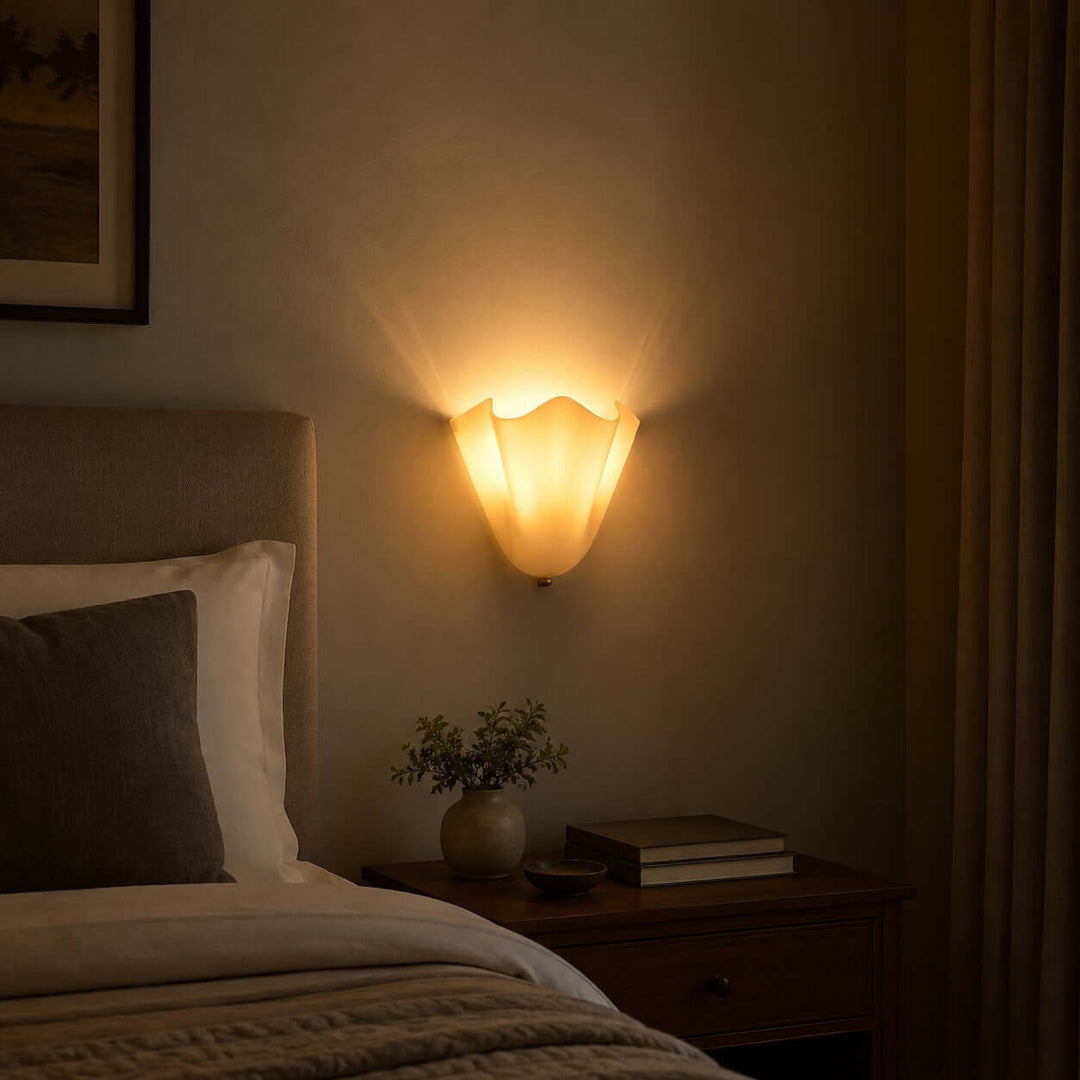 Beige glass wall sconce casting warm light beside a bed at night.