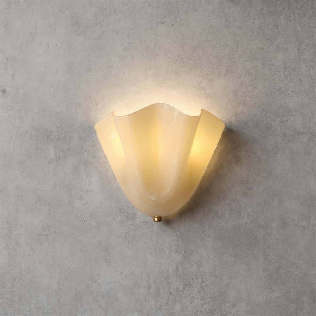 Beige glass wall sconce with gold hardware glowing on a gray wall.