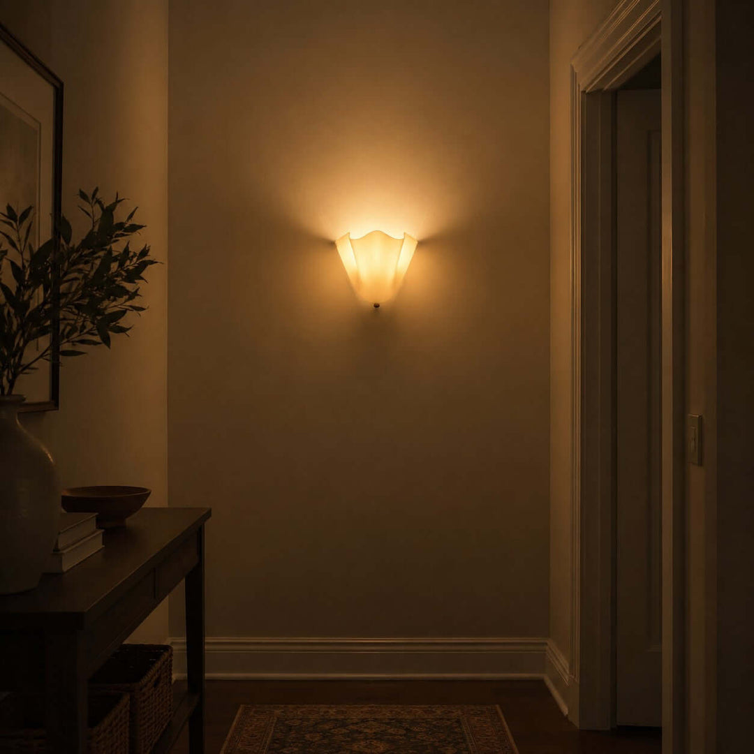 Beige glass wall sconce glowing on a hallway wall in a dim interior.