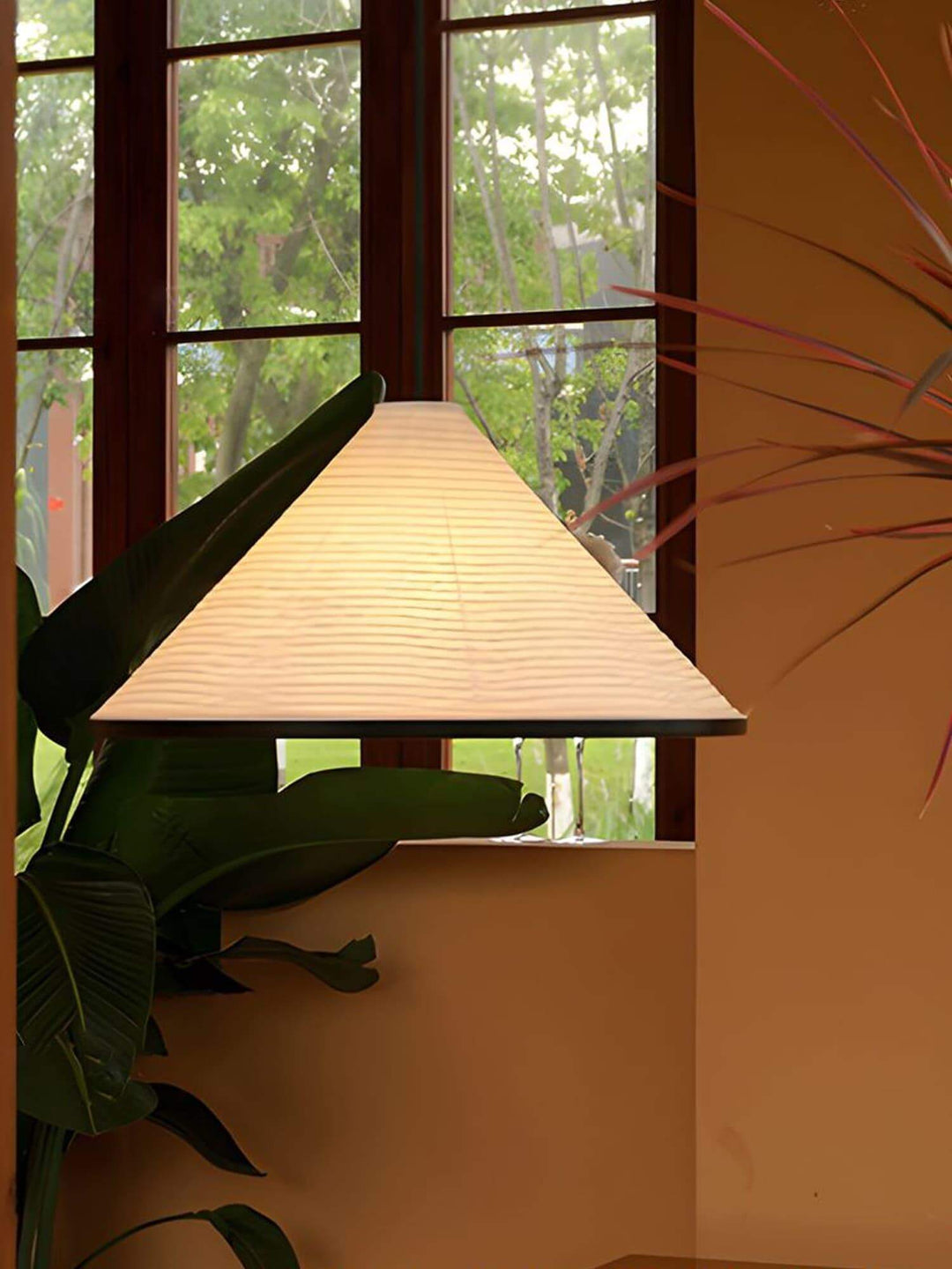 Close view of the glowing Washi Paper Hat Pendant Lamp beside a wood-framed window and tropical plant