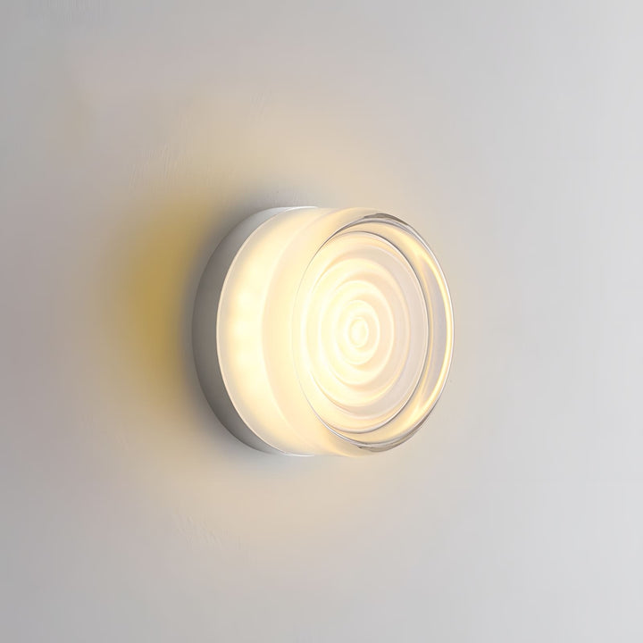 White glass wall sconce with concentric front texture glowing on a gray wall.